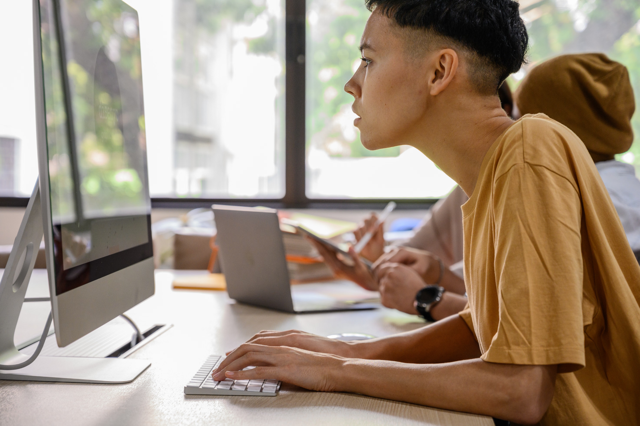 A young man working on a desktop computer while typing on a keyboard in a bright, modern office, with a colleague working in the background.